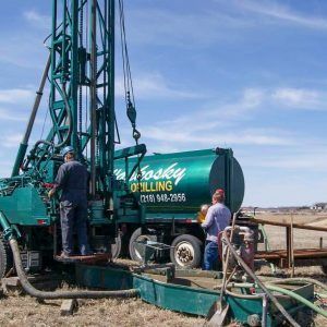 Two Waskosky Well Drilling employees work at a green piece of Waskosky Well Drilling equipment in a field.